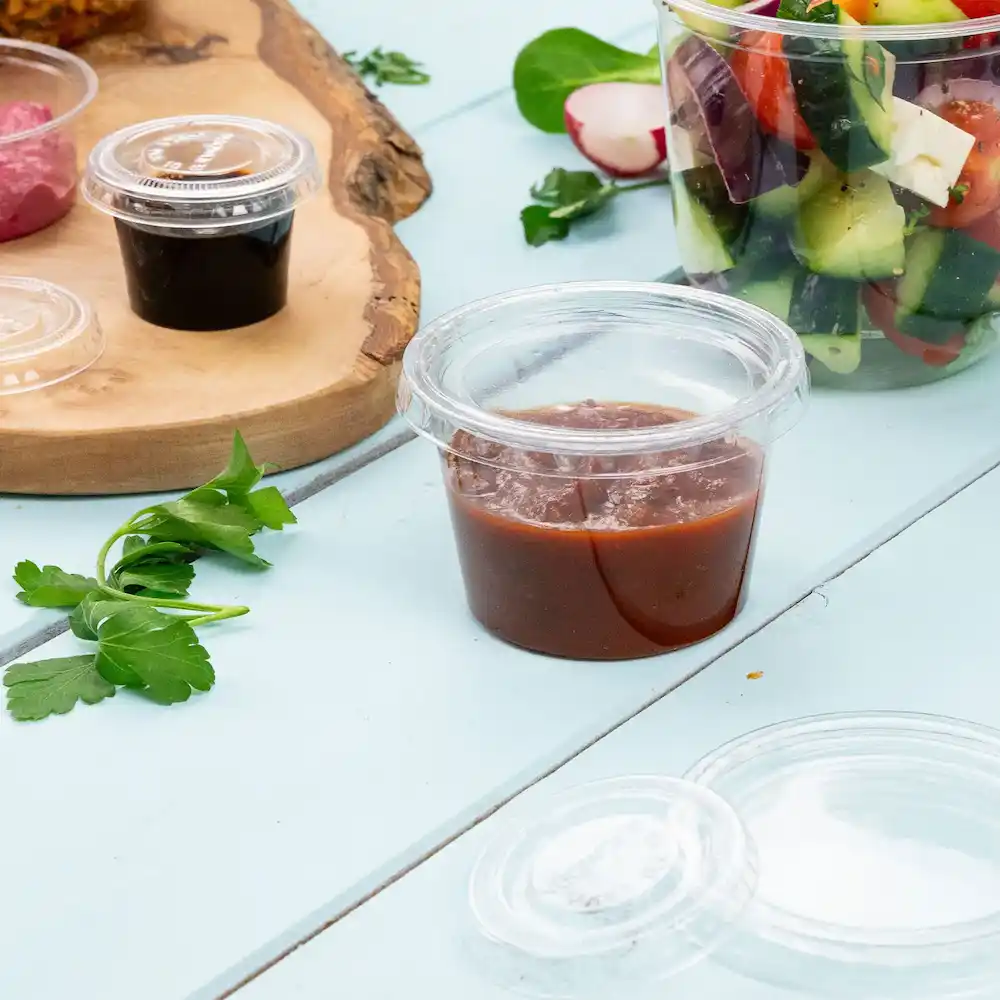 Contenant transparent avec une sauce rougeâtre, accompagné d'herbes vertes et d'autres aliments sur une surface bleu clair.