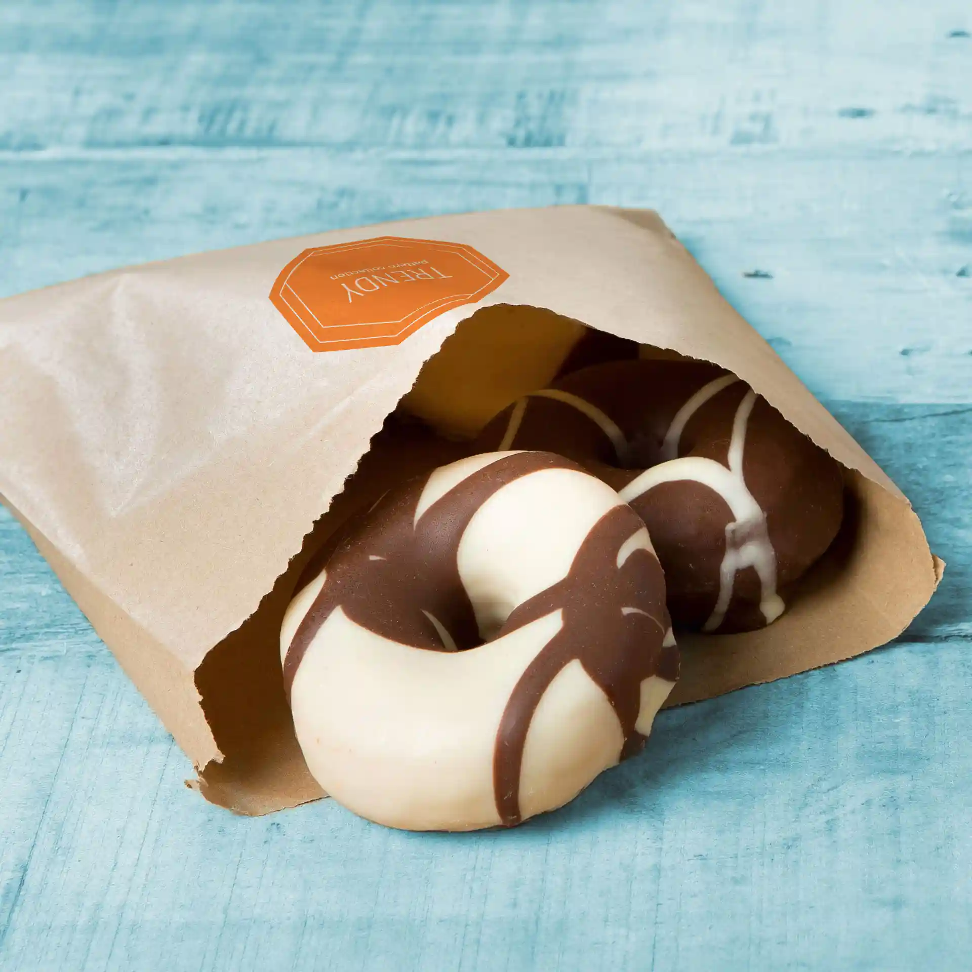Sac en papier brun plat avec des donuts enrobés de chocolat partiellement visibles à l'intérieur, posé sur une surface en bois bleu.