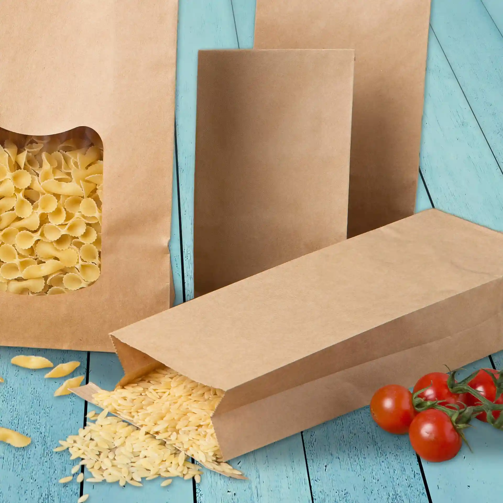 Sacs en papier kraft marron, un sac ouvert renversant des pâtes, à côté de deux sacs debout sur une surface en bois bleu.