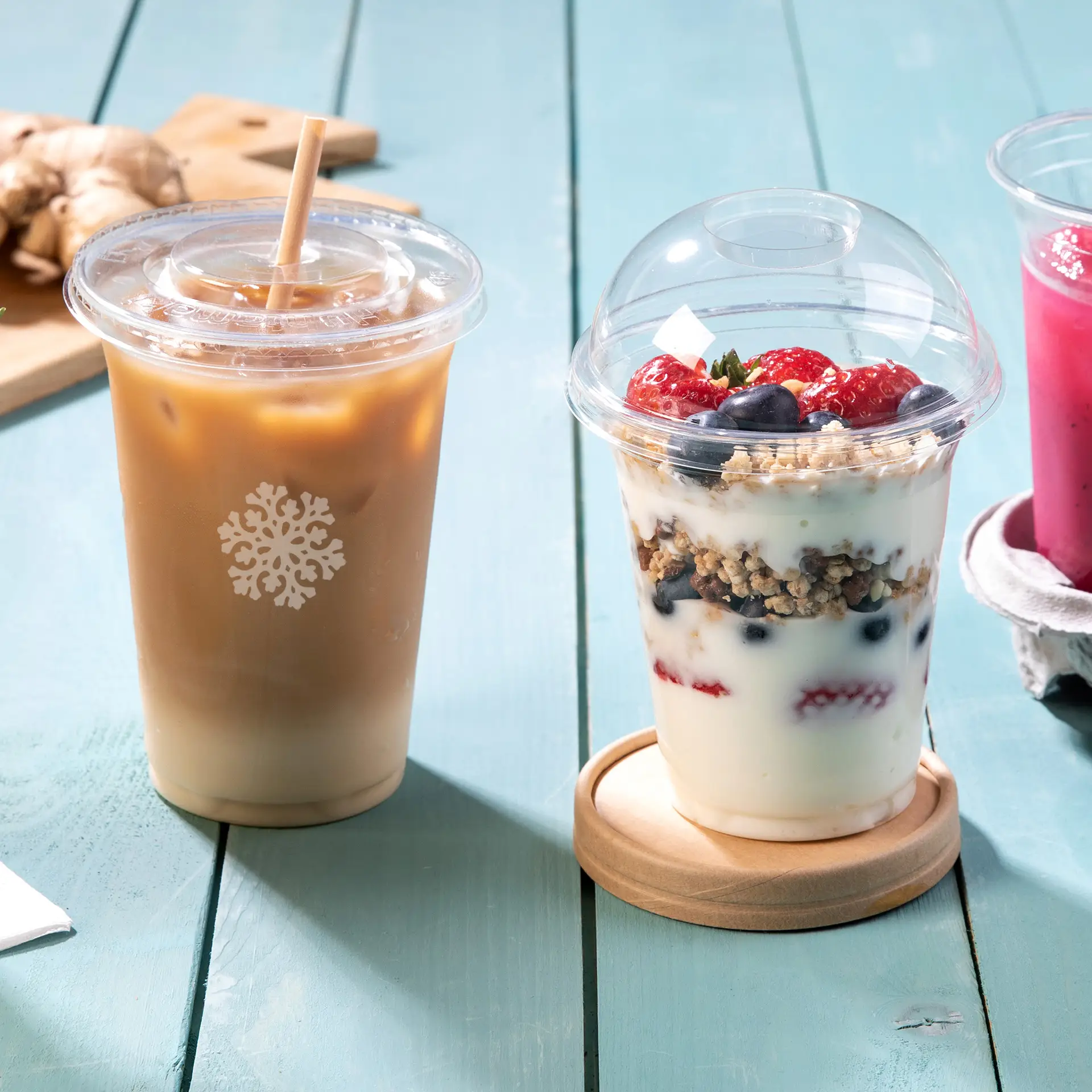 Trois gobelets PLA transparents pour boissons froides : un gobelet de café glacé, un dessert aux baies et une boisson rose, posés sur une table en bois.