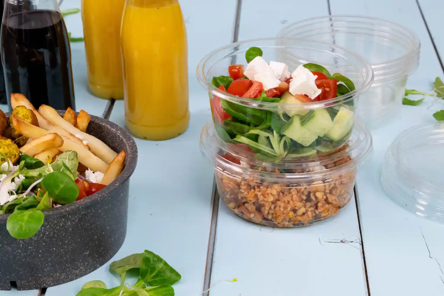 Contenants contenant une salade et des céréales à côté d'un bol de frites ; deux bouteilles de boisson en arrière-plan ; surface en bois bleu clair.