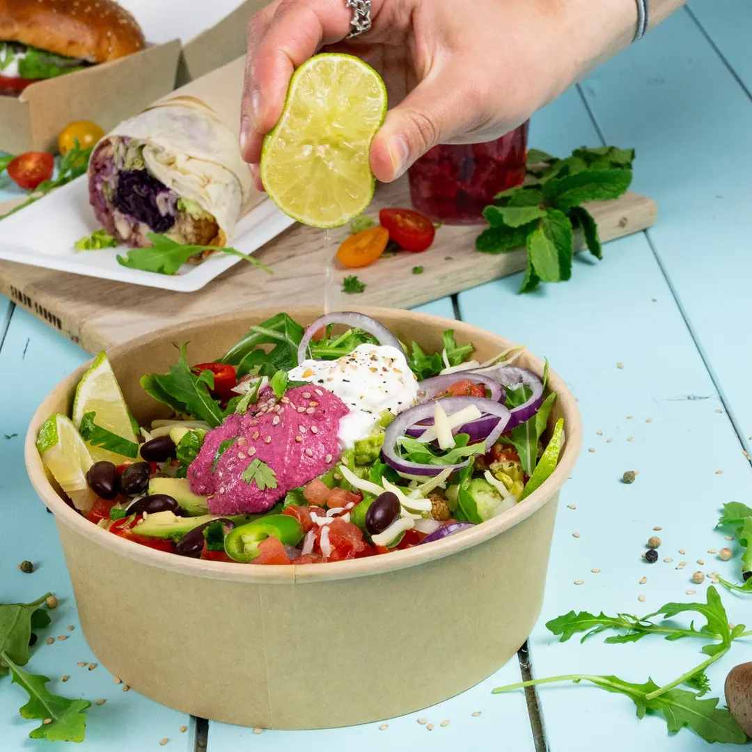 Une main presse un citron vert sur une salade dans un bol marron, avec des légumes verts, des tomates, des oignons et du houmous rose.