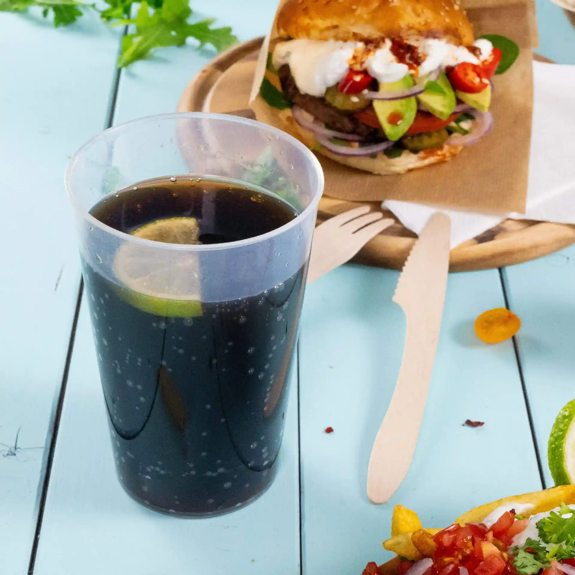 Gobelet réutilisable transparent rempli d'une boisson sombre et d'une tranche de citron vert, placé à côté d'un burger et d'ustensiles en bois.