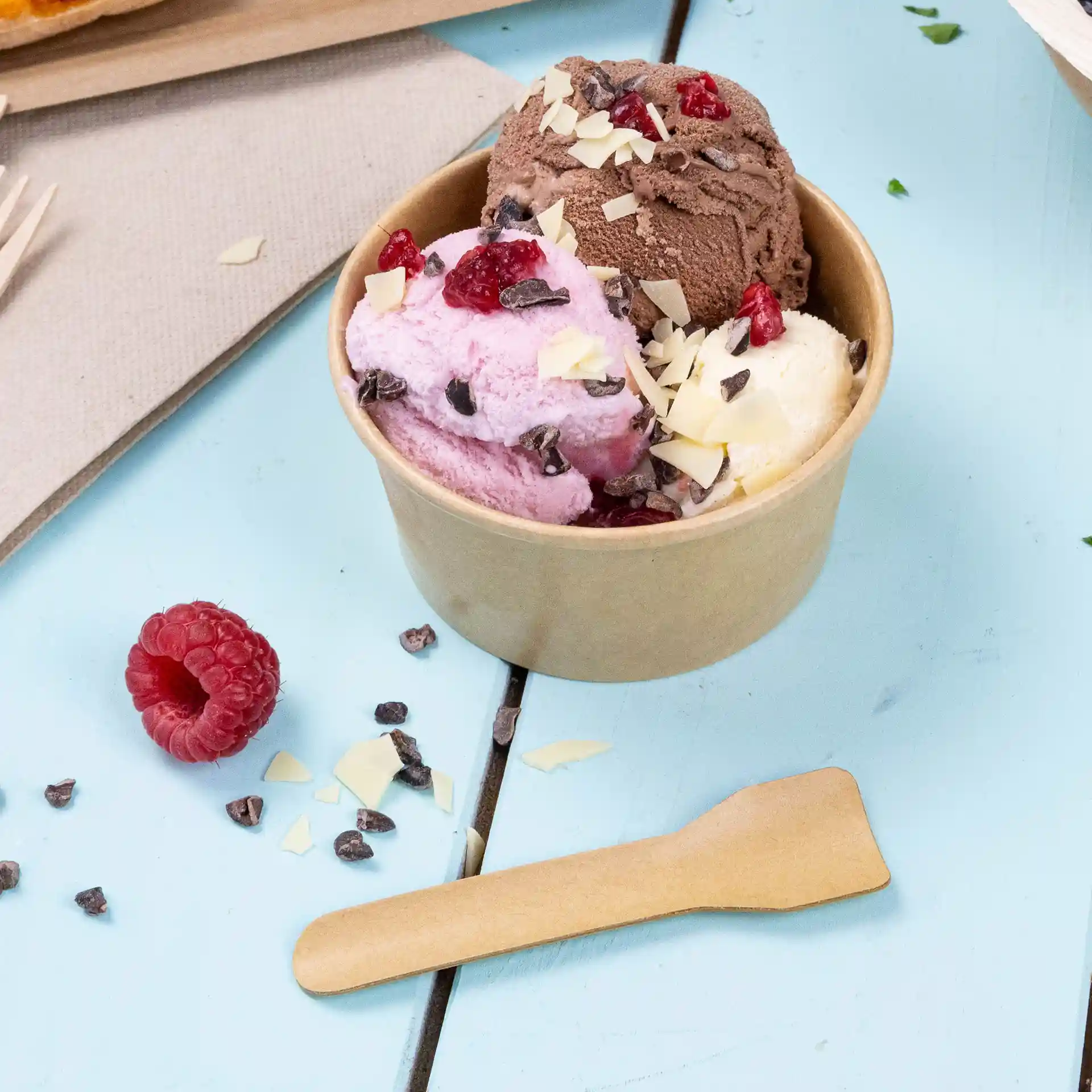 Gobelet contenant trois boules de glace rose, brune et blanche, surmonté de pépites de chocolat et de framboises, avec une cuillère en bois.