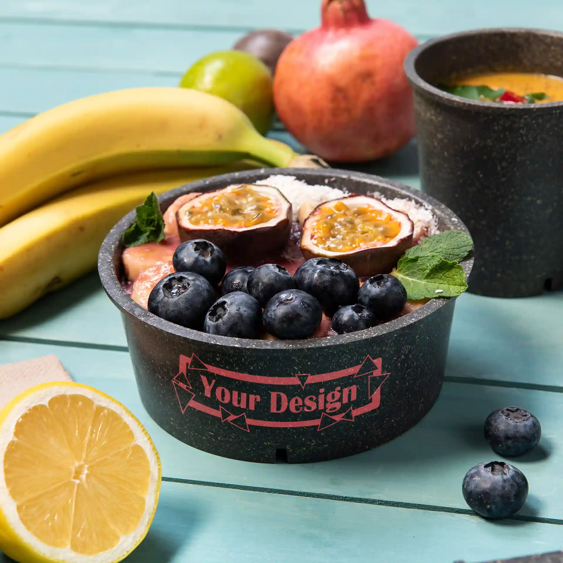 Bol rond gris réutilisable avec des garnitures de fruits, y compris des myrtilles et des fruits de la passion, sur une surface turquoise.