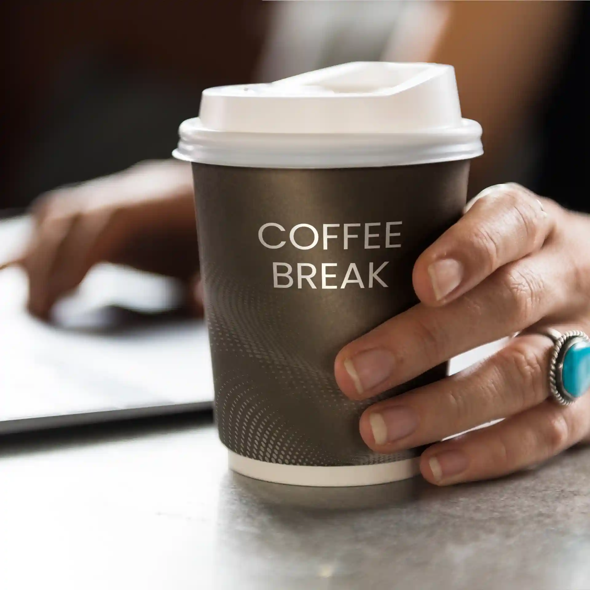 Gobelet en papier brun avec un couvercle blanc, tenu dans une main, portant le texte "PAUSE CAFÉ".