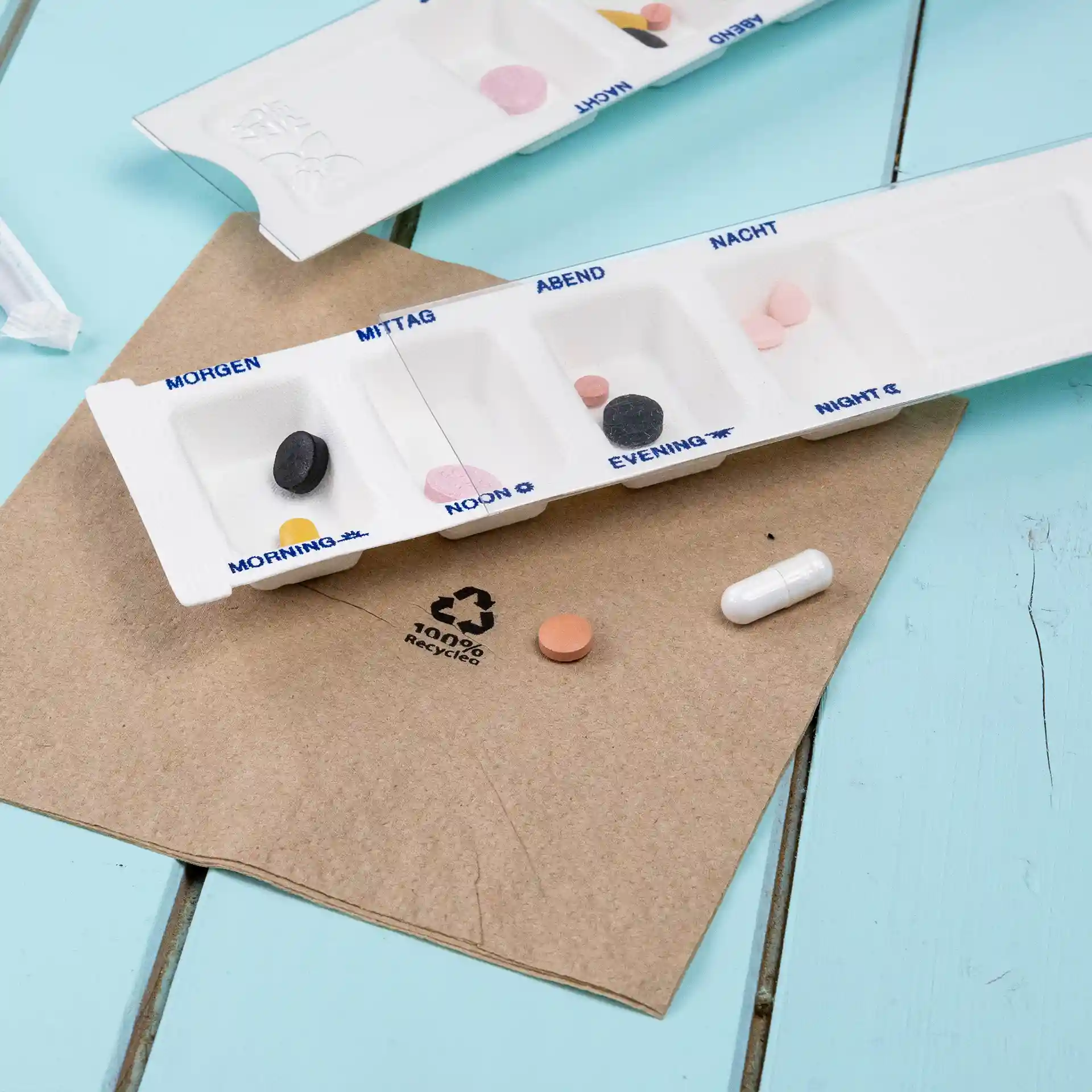 Distributeur de médicaments à quatre compartiments, blanc, sur une surface brune avec des pilules colorées à proximité.