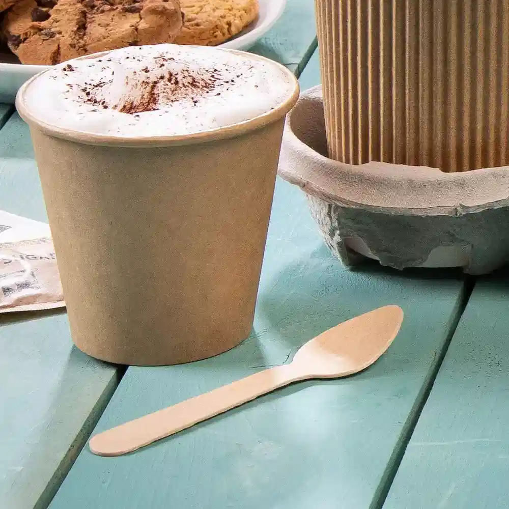 Gobelet en carton marron avec une couche de mousse et une cuillère en bois sur une table turquoise, avec des biscuits en arrière-plan.