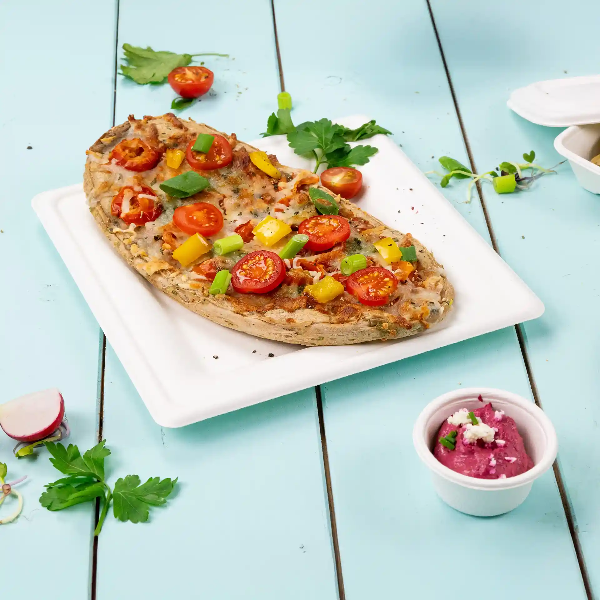 Assiette carrée blanche en canne à sucre avec une pizza garnie de tomates, poivrons et oignons verts, accompagnée d'un petit pot de sauce.
