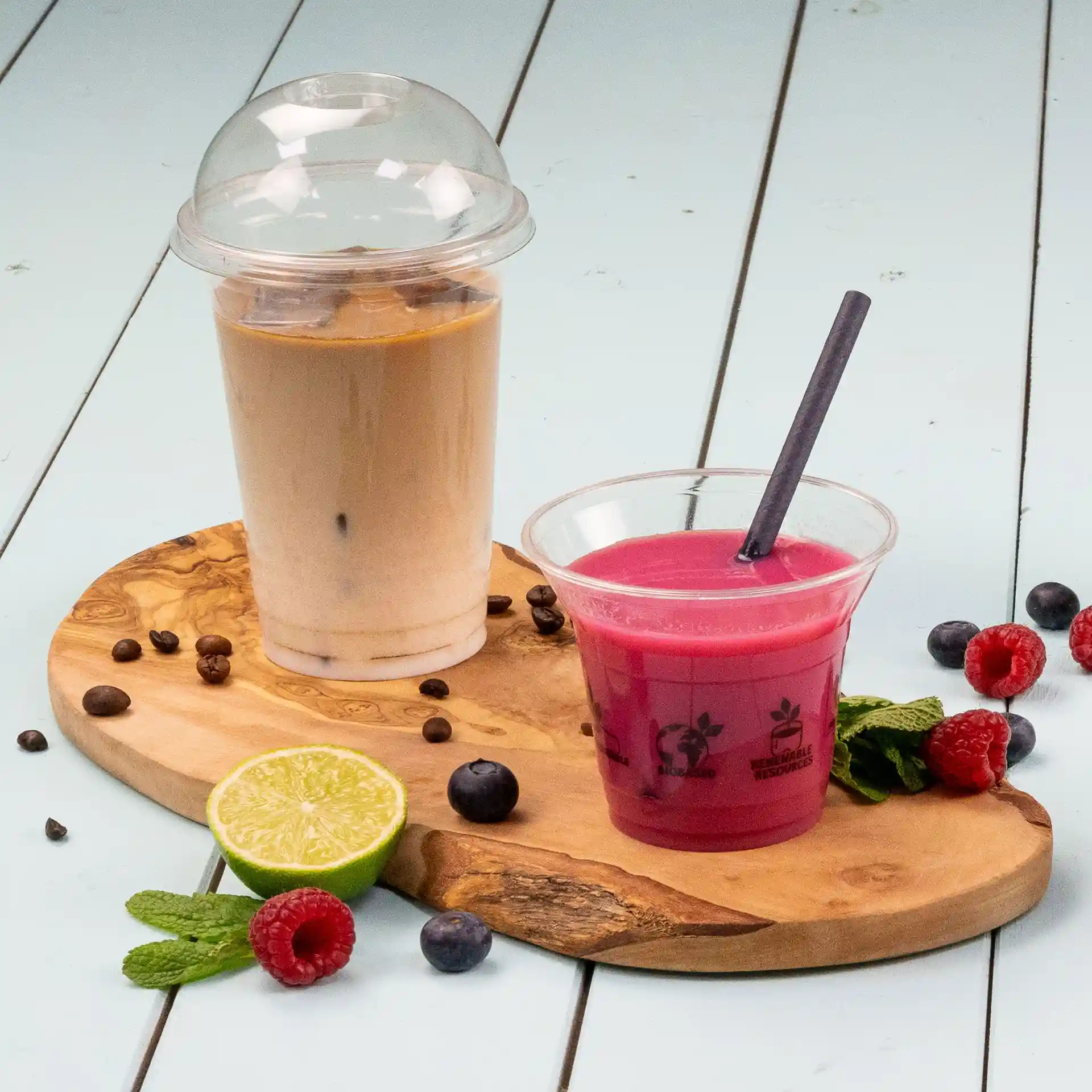 Image d'un gobelet transparent avec un couvercle en dôme contenant une boisson brun clair, à côté d'une boisson rose dans un gobelet transparent avec une paille, sur une planche en bois avec du citron vert, des baies et de la menthe.