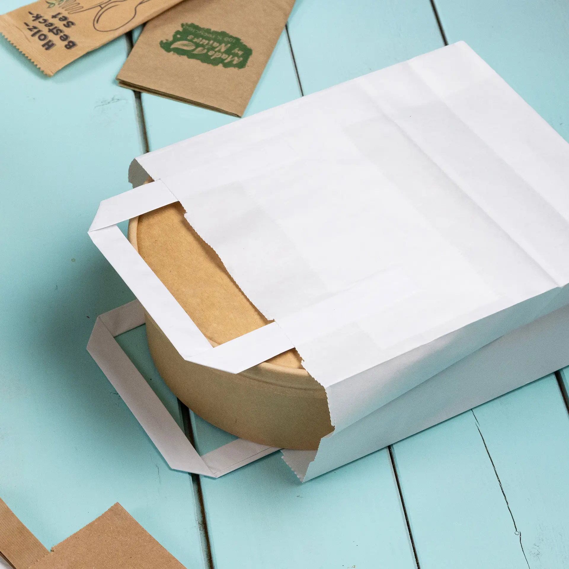 Sac à papier blanc partiellement ouvert, révélant un conteneur en kraft rond à l'intérieur sur une surface bleu clair.