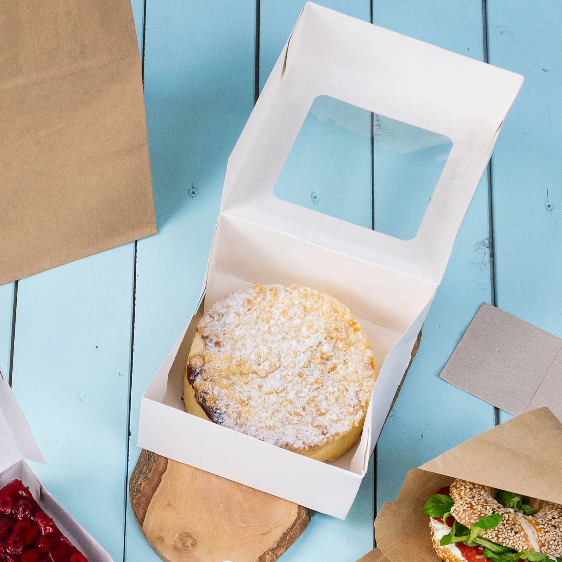 Boîte à gâteaux M blanche avec un couvercle ouvert montrant un gâteau rond à l'intérieur, placé sur une surface en bois avec d'autres aliments à proximité.