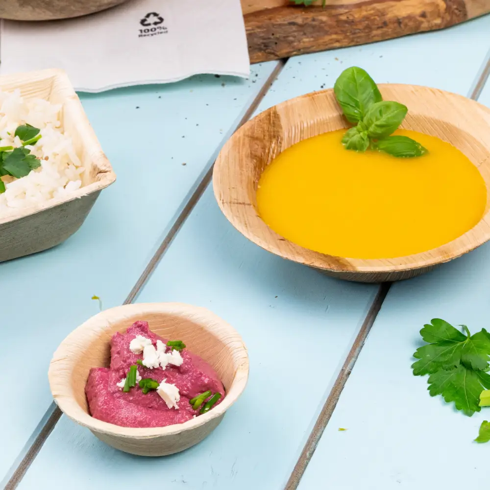 Bol en bambou rempli de soupe jaune garni d'une feuille de basilic ; petit bol avec une trempette rose surmontée d'herbes ; riz aux herbes dans un contenant à proximité.