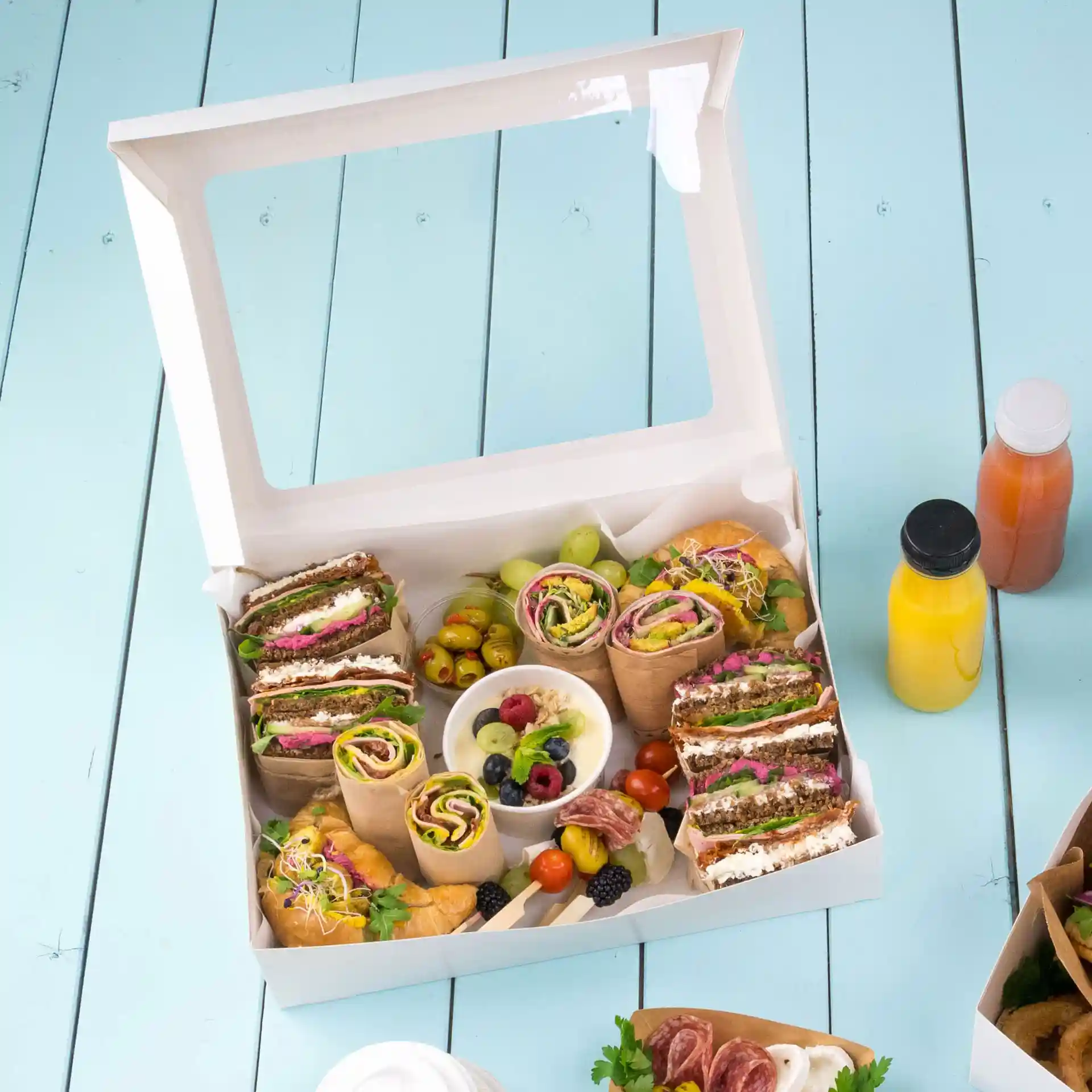 Boîte contenant des wraps assortis, des sandwiches, des fruits et un petit bol de baies mélangées, placée sur une surface bleu clair.