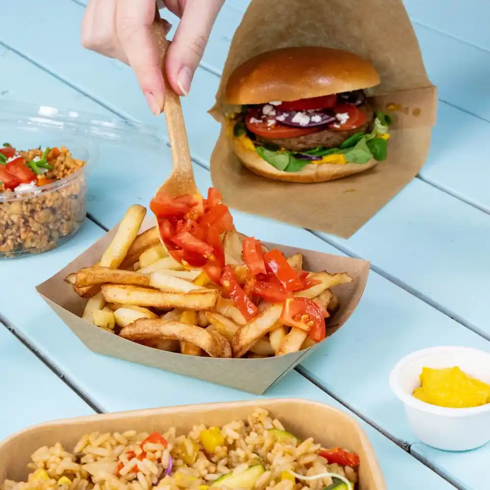 Une main tenant une cuillère en bois ajoute des tomates dés à une portion de frites dans un plateau marron ; un burger et un gobelet de riz sont en arrière-plan.