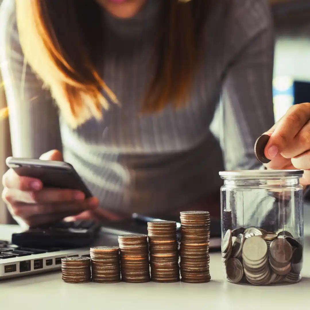 Femme mettant une pièce dans un bocal en verre tout en utilisant un smartphone, avec des piles de pièces sur une table.