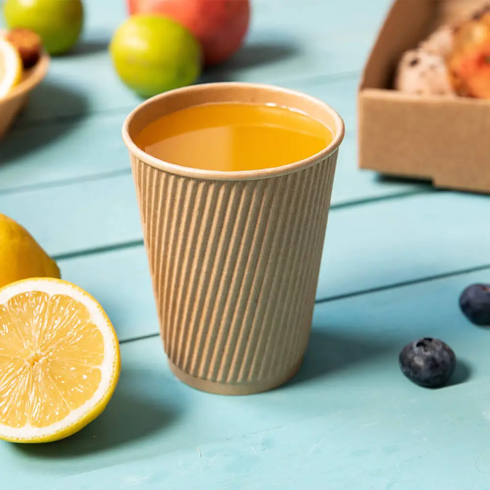 Un gobelet en carton cannelé rempli d'un liquide jaune, entouré de tranches de citron, de fruits et d'une boîte de pâtisseries.