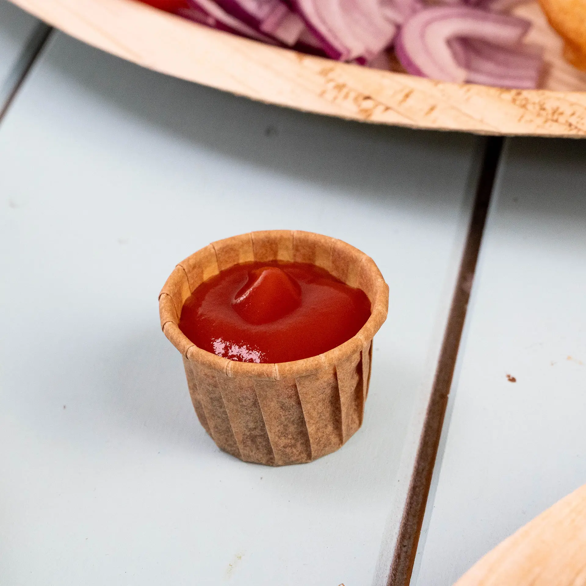 Gobelet en papier brun rempli de ketchup sur une surface bleu clair, avec des oignons rouges flous en arrière-plan.