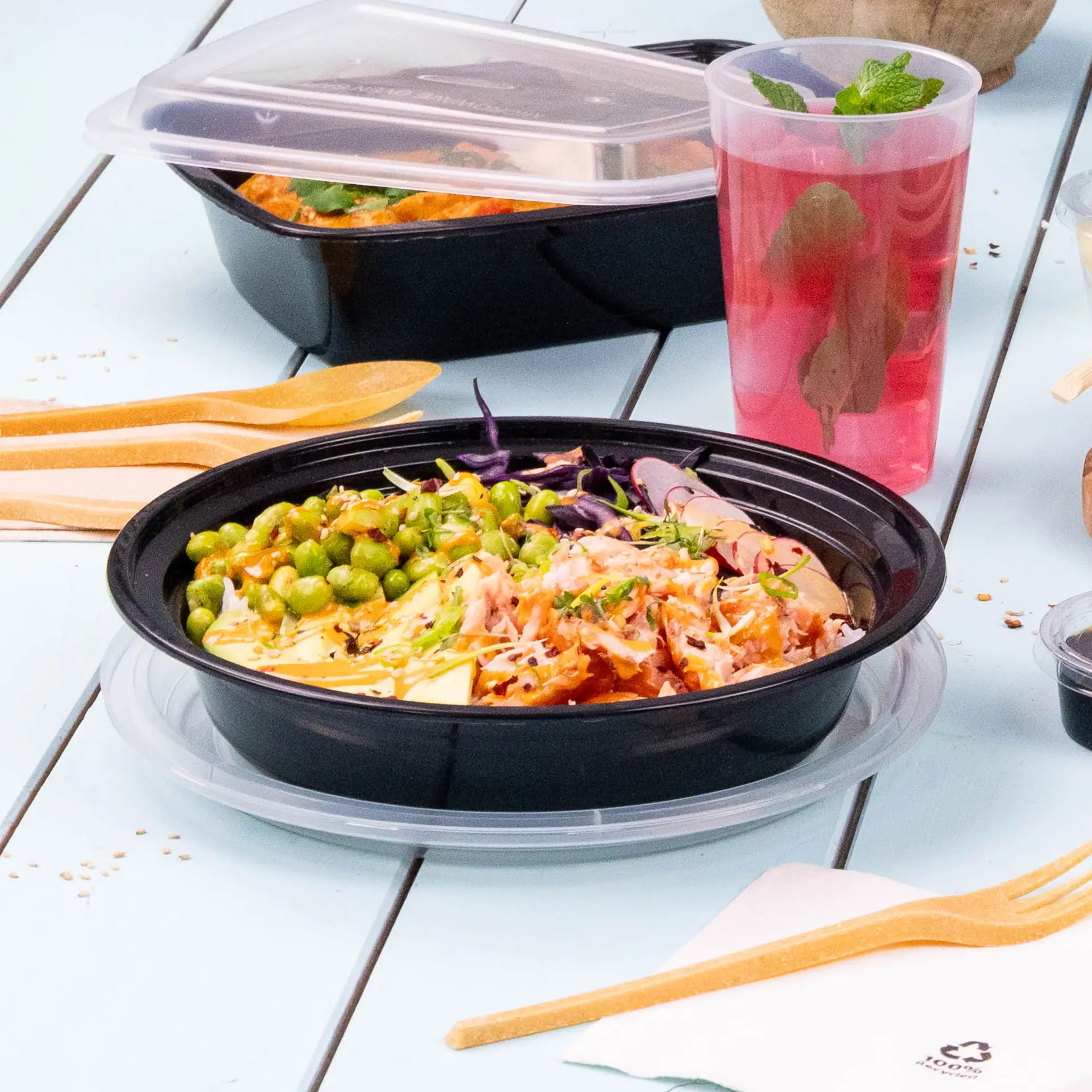 Assiette noire réutilisable pour plats chauds et froids, servie avec des petits pois, des légumes râpés et une boisson ; comprend des ustensiles en bois.