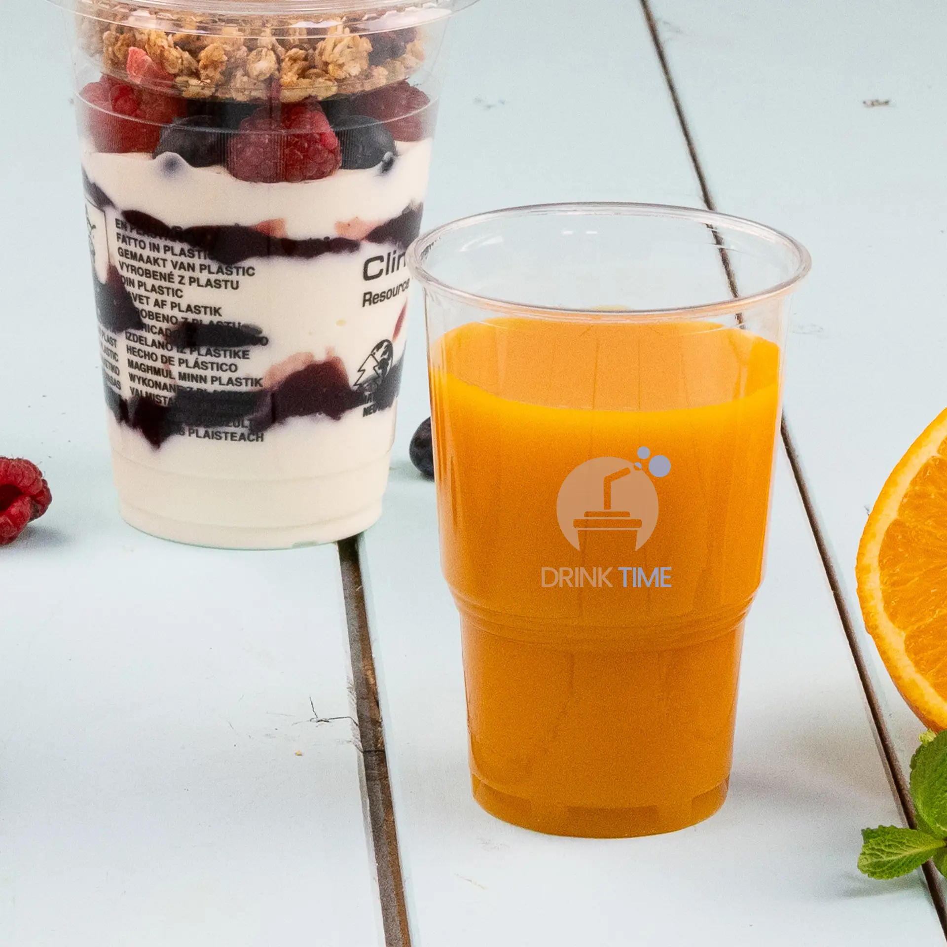 Un gobelet rPET transparent rempli de jus d'orange, avec un logo en forme de diamant bleu ; accompagné d'une orange fraîche et de feuilles de menthe à proximité.