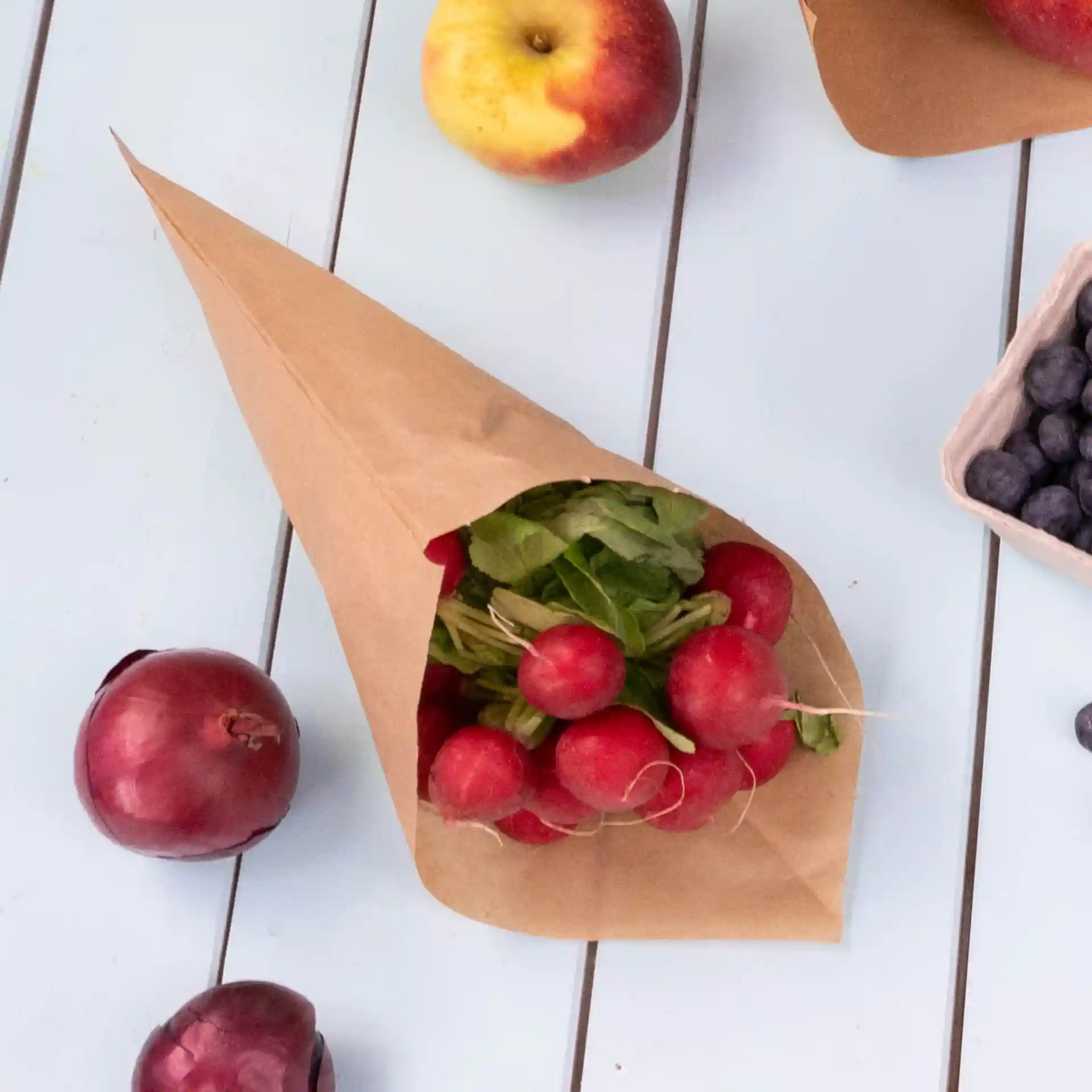 Sac en papier brun rempli de radis rouges et de légumes verts, avec des pommes et des myrtilles à proximité sur une surface bleu clair.