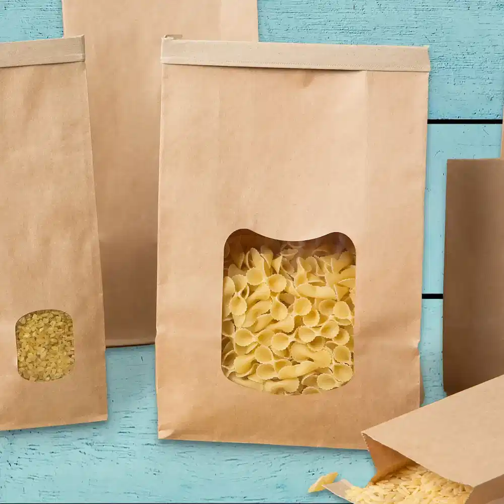 Sacs en papier marron avec des fenêtres transparentes, remplis de différentes formes de pâtes, sur une surface en bois bleu.