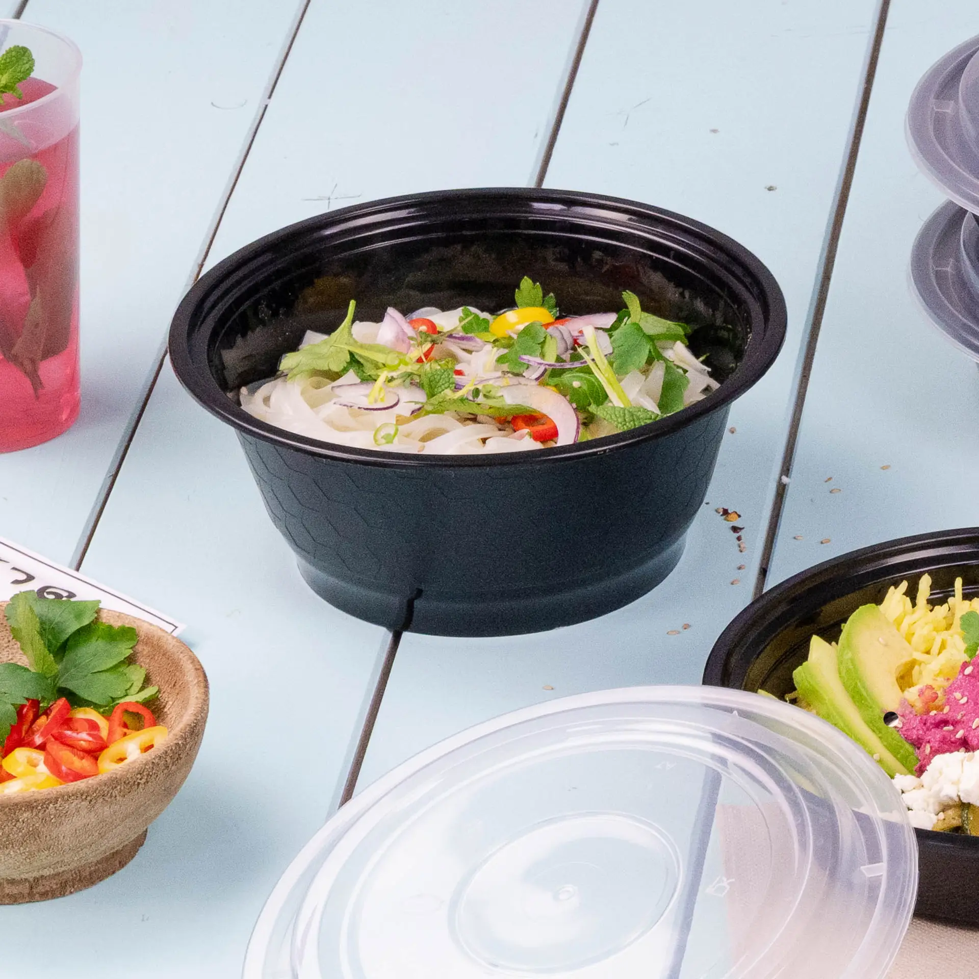 Un récipient alimentaire réutilisable noir rempli de nouilles, garni d'herbes et de piments, entouré d'autres aliments.