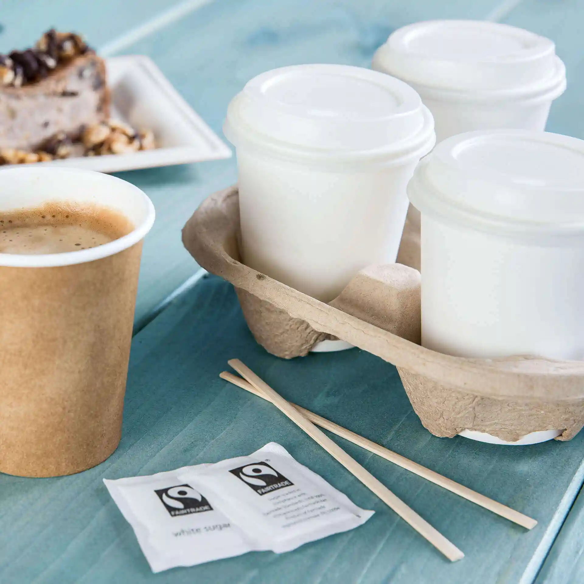 Image d'un gobelet en papier brun avec du café, trois gobelets blancs dans un support, des bâtonnets en bois et des sachets de sucre sur une surface bleue.