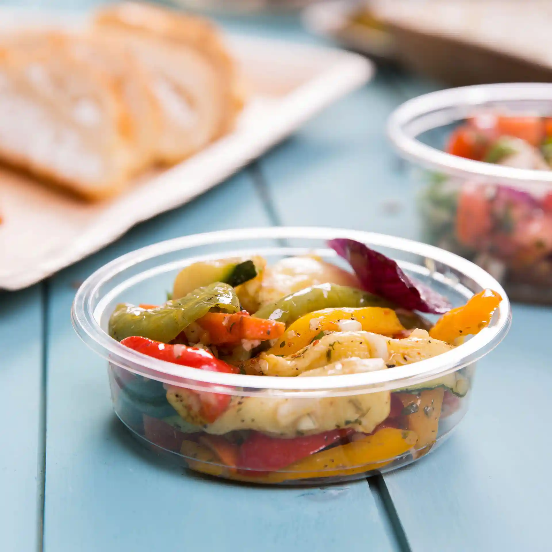 Conteneur rond transparent pour deli avec légumes mélangés, capacité de 250 ml, sur une surface bleue avec du pain en arrière-plan.