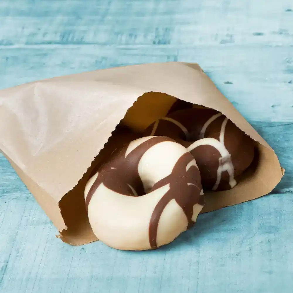 Des beignets enrobés de chocolat partiellement visibles dans un sac en papier brun sur une surface en bois bleu.
