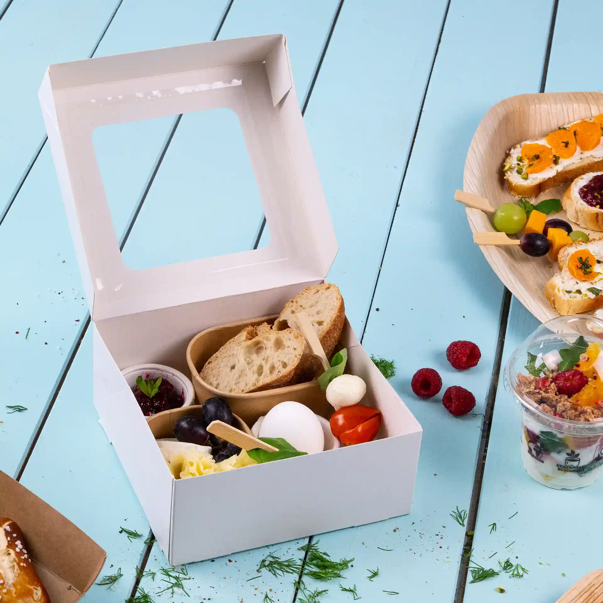Boîte en carton blanche avec une fenêtre transparente, contenant du pain, du fromage, un œuf et des fruits assortis.