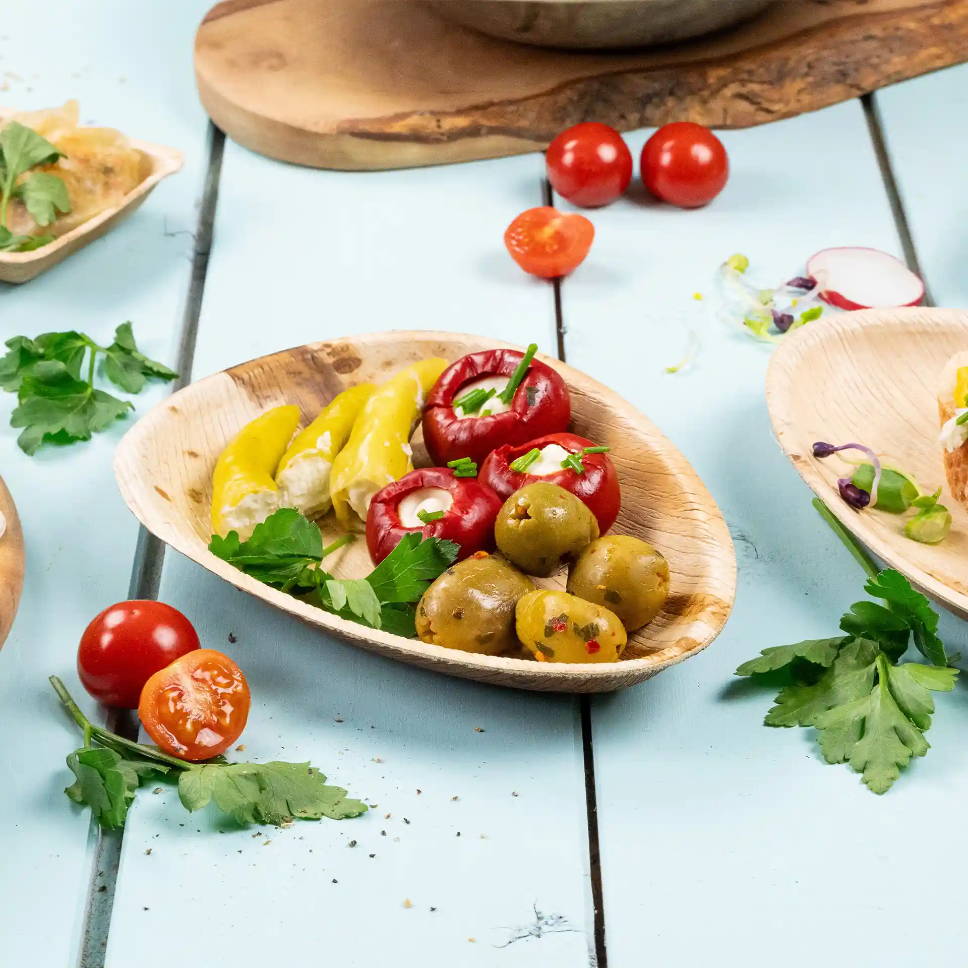 Assiette ovale en feuille de palmier avec des poivrons jaunes, des tomates farcies rouges, des olives vertes et du persil sur une surface bleue.