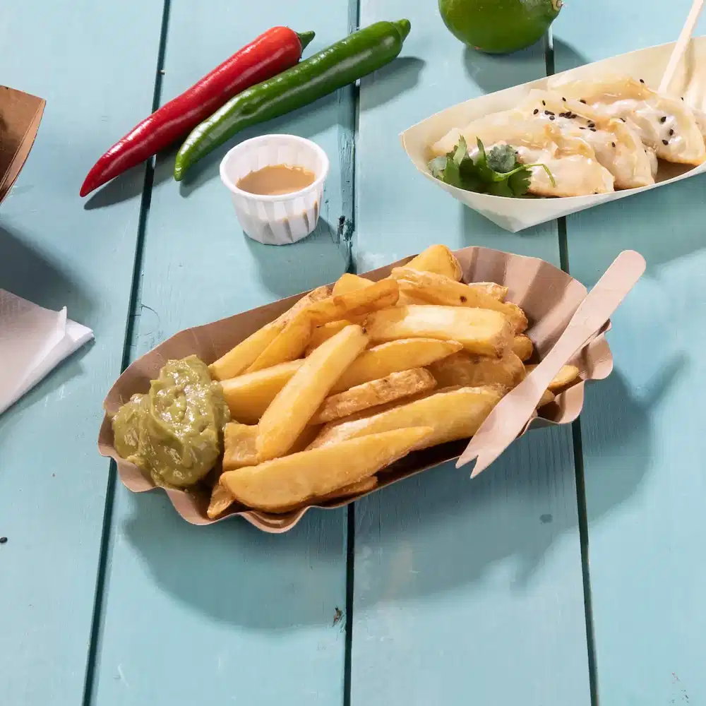 Panier de frites avec sauce verte, ustensiles en bois et piments sur une table en bois bleue.