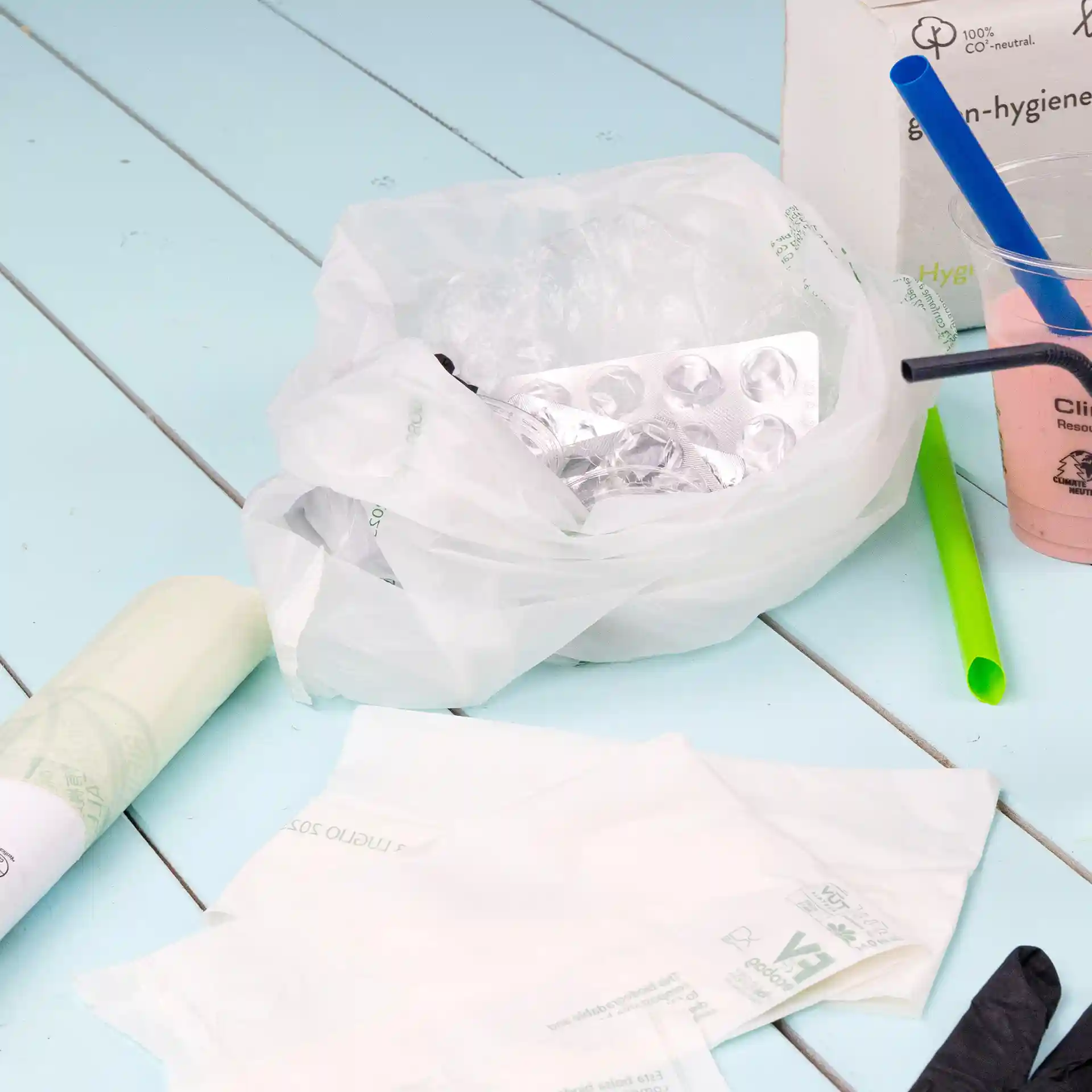 Sac poubelle bio transparent avec contenu froissé, y compris un emballage blister, sur une surface bleu clair.