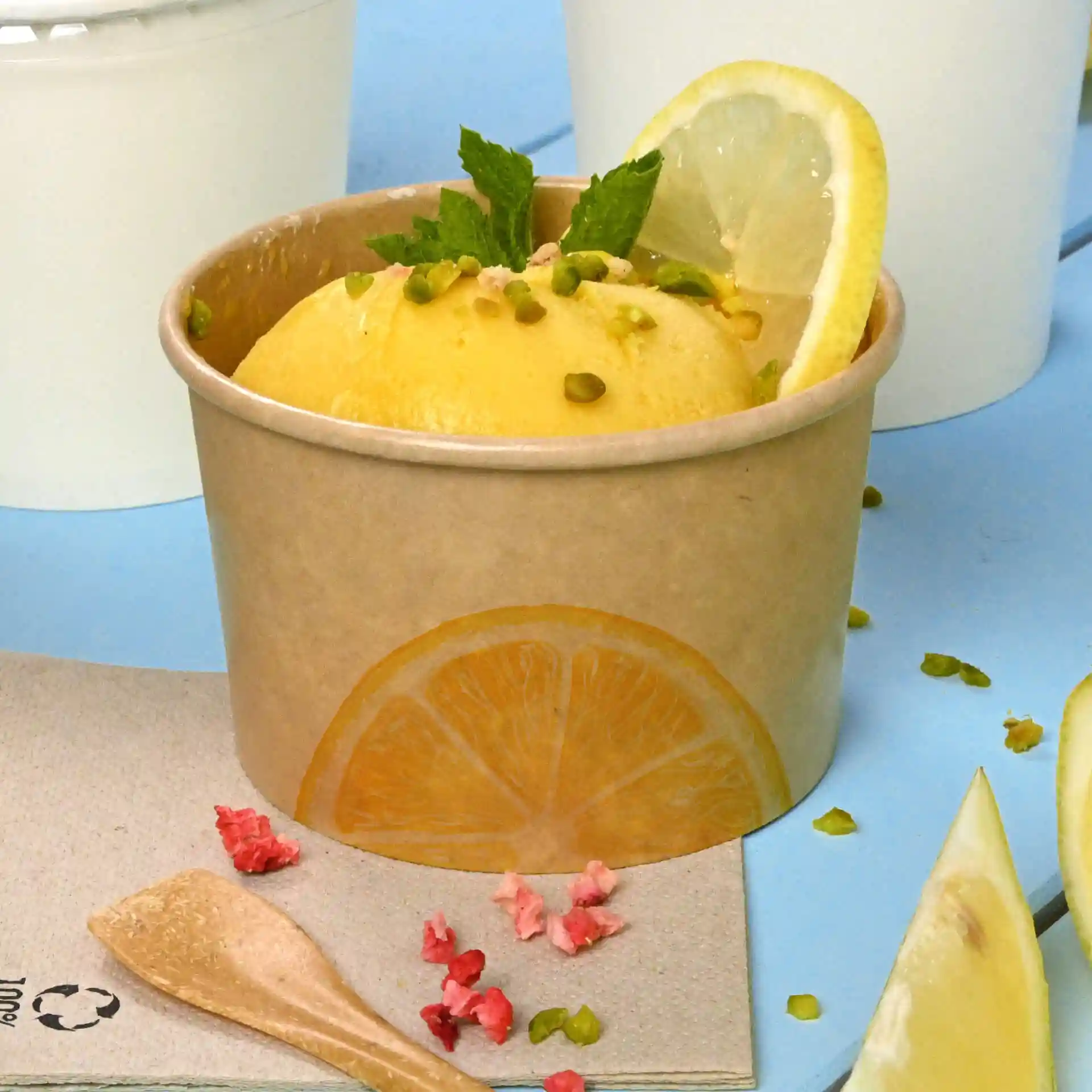Gobelet en carton brun avec sorbet au citron, garni de feuilles de menthe et de pistaches, sur fond bleu.