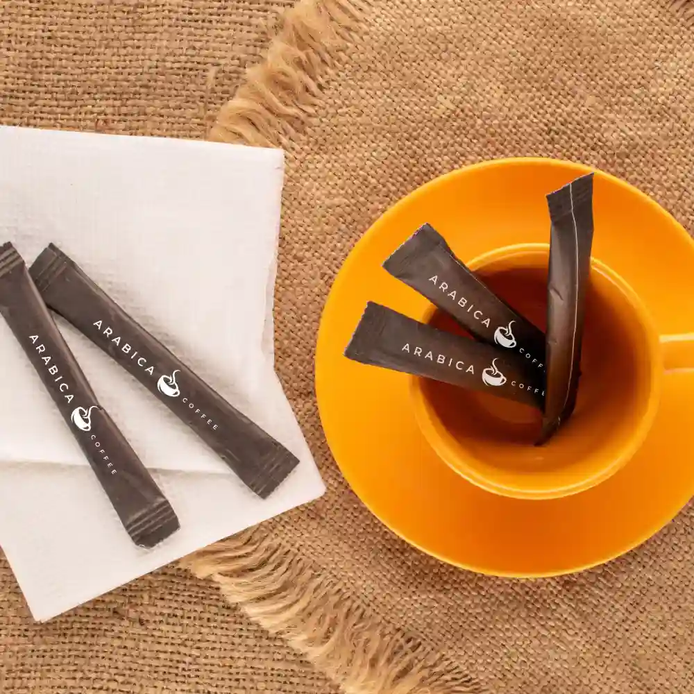 Plusieurs sachets de sucre Arabica à côté d'une serviette blanche sur une surface en jute et dans une tasse à café orange.