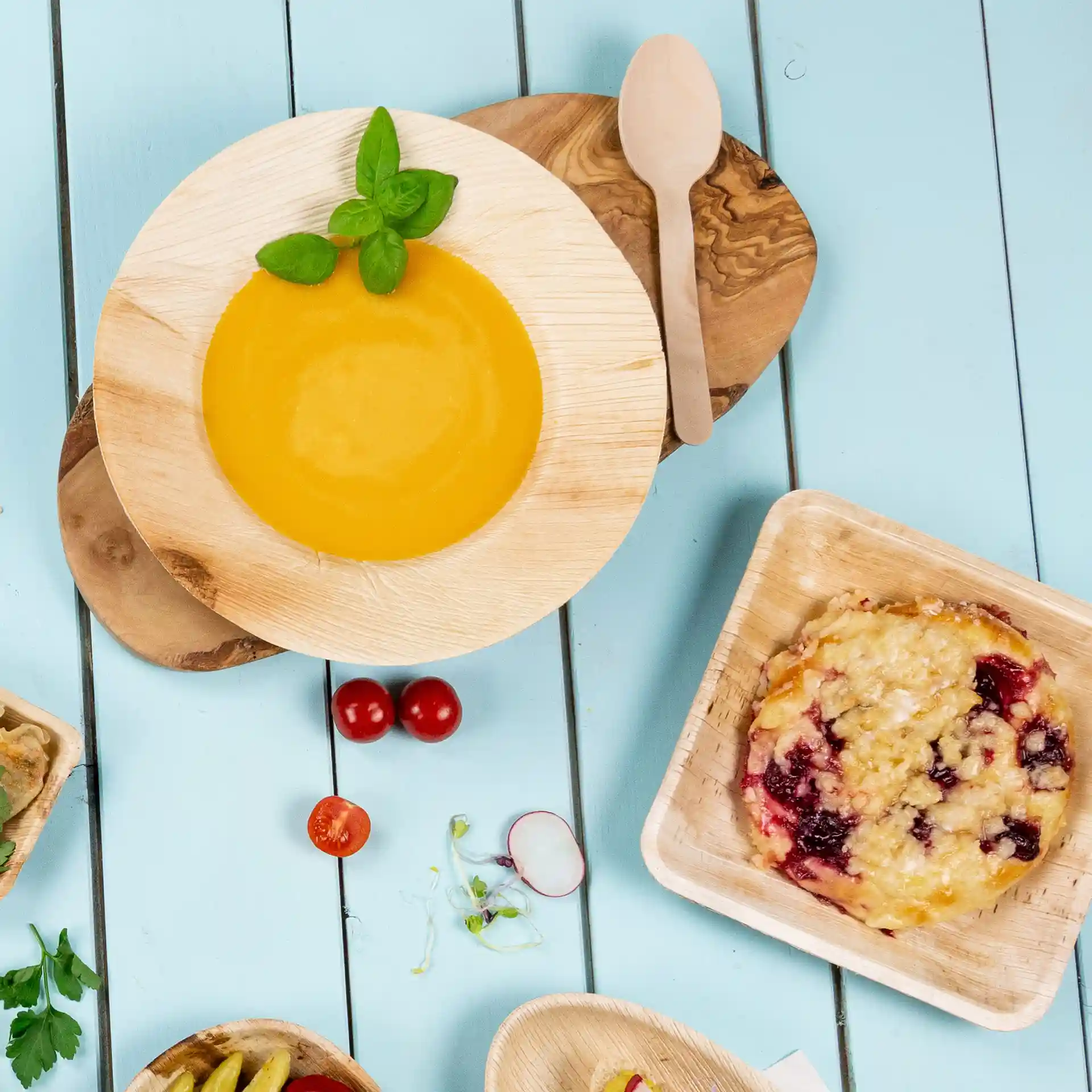 Assiette ronde profonde en feuille de palmier avec de la soupe jaune, garnie de basilic, et une cuillère en bois sur une surface en bois bleu.