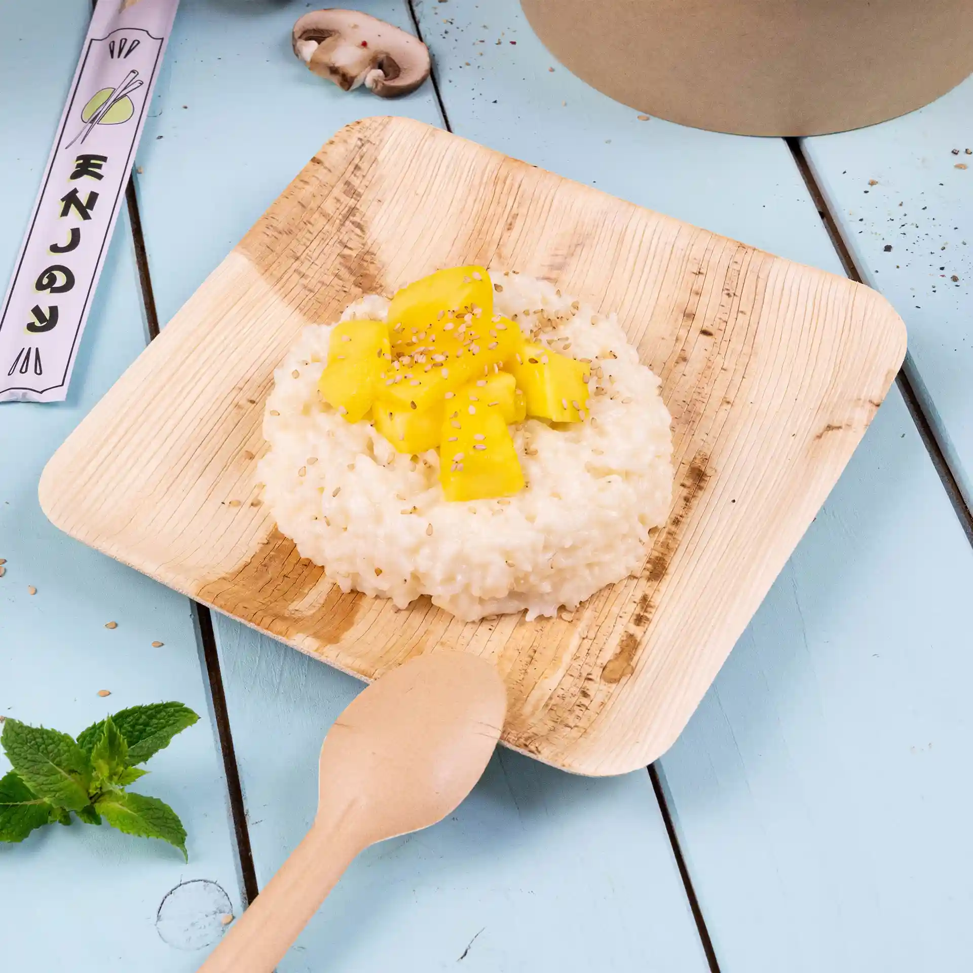 Assiette carrée en feuille de palmier avec risotto garni de mangue et de graines de sésame, accompagnée d'une cuillère en bois sur une surface bleue.