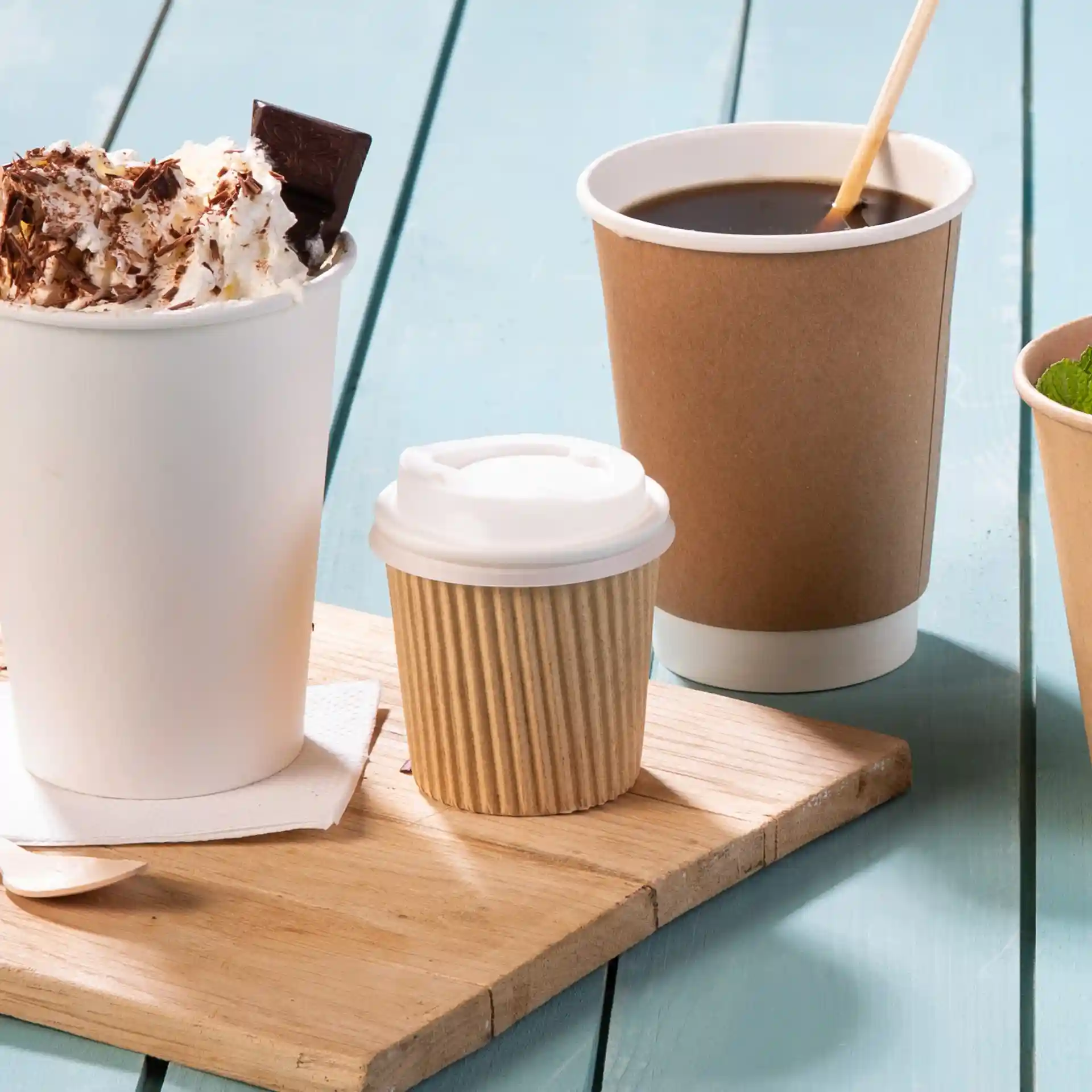 Image d'une planche en bois avec une grande tasse blanche surmontée de crème fouettée et de chocolat, une petite tasse brune striée et une tasse à café brune.
