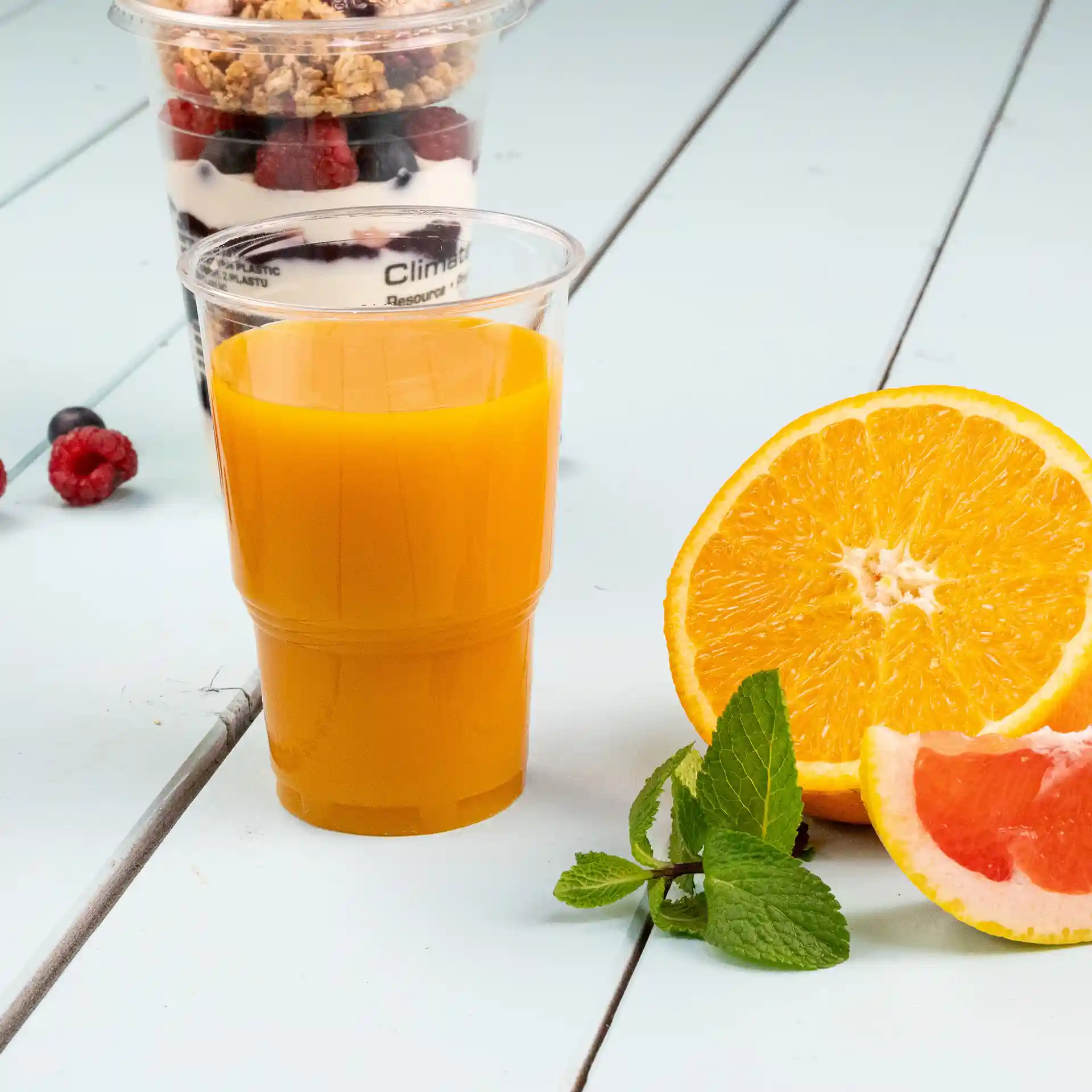 Gobelet en plastique transparent rempli de jus d'orange, avec une orange coupée en deux et des feuilles de menthe à côté.