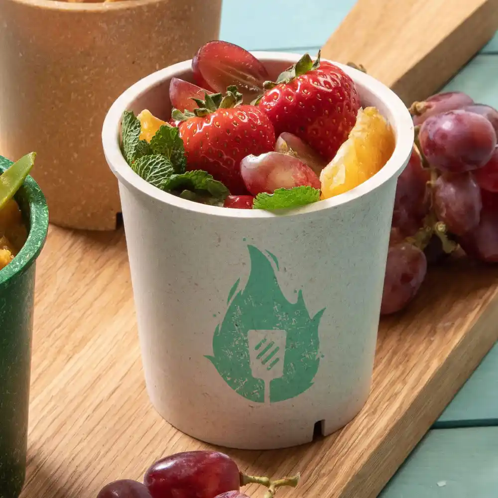 Tasse blanche remplie de fruits mélangés, avec un logo de flamme verte, à côté d'une tasse verte et de raisins sur une surface en bois.