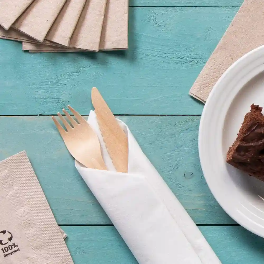 Ustensiles en bois enveloppés dans une serviette blanche, accompagnés de serviettes en papier recyclé et d'un morceau de gâteau sur une assiette.