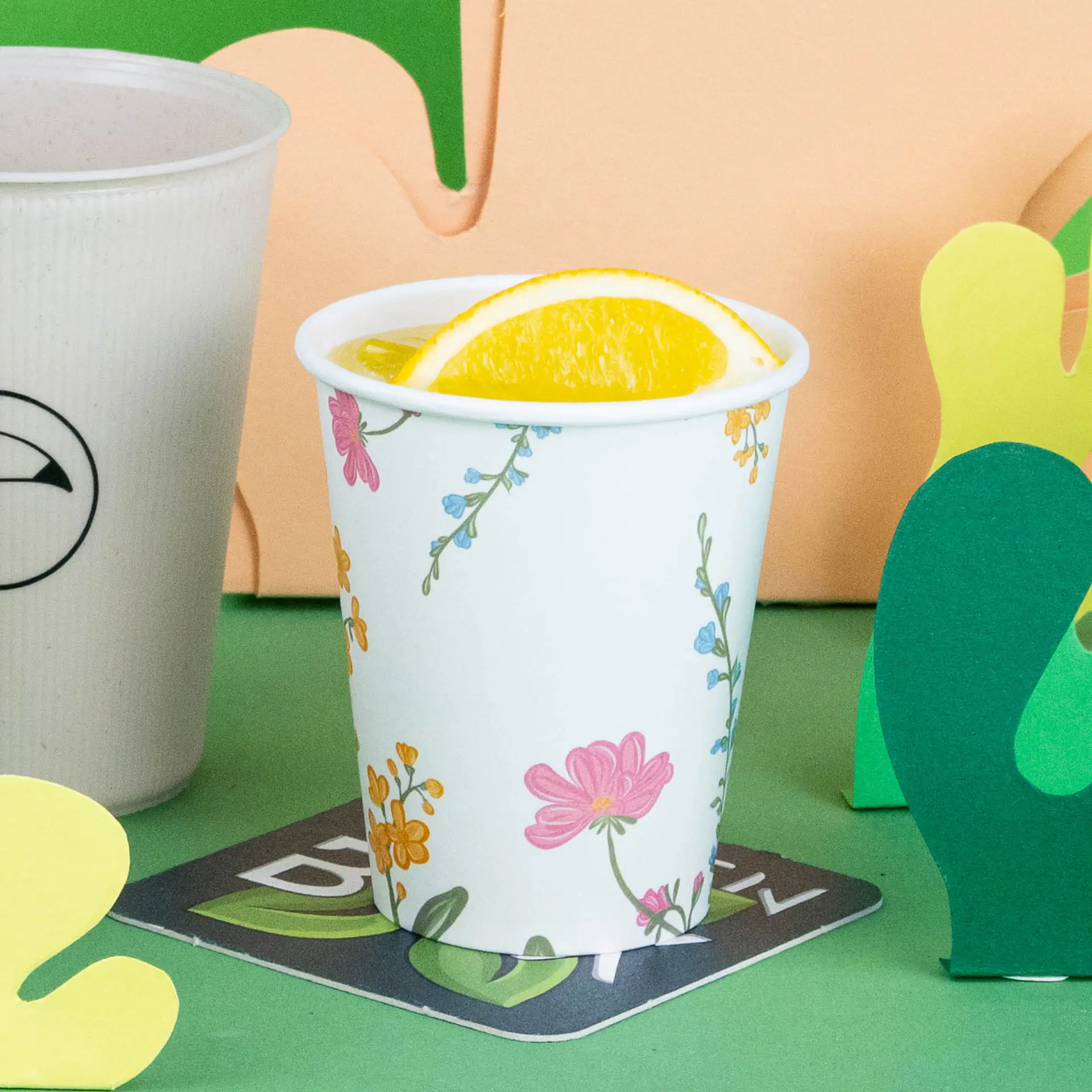 Gobelet blanc avec un design floral, rempli d'une boisson et d'une tranche d'orange, sur un fond vert.