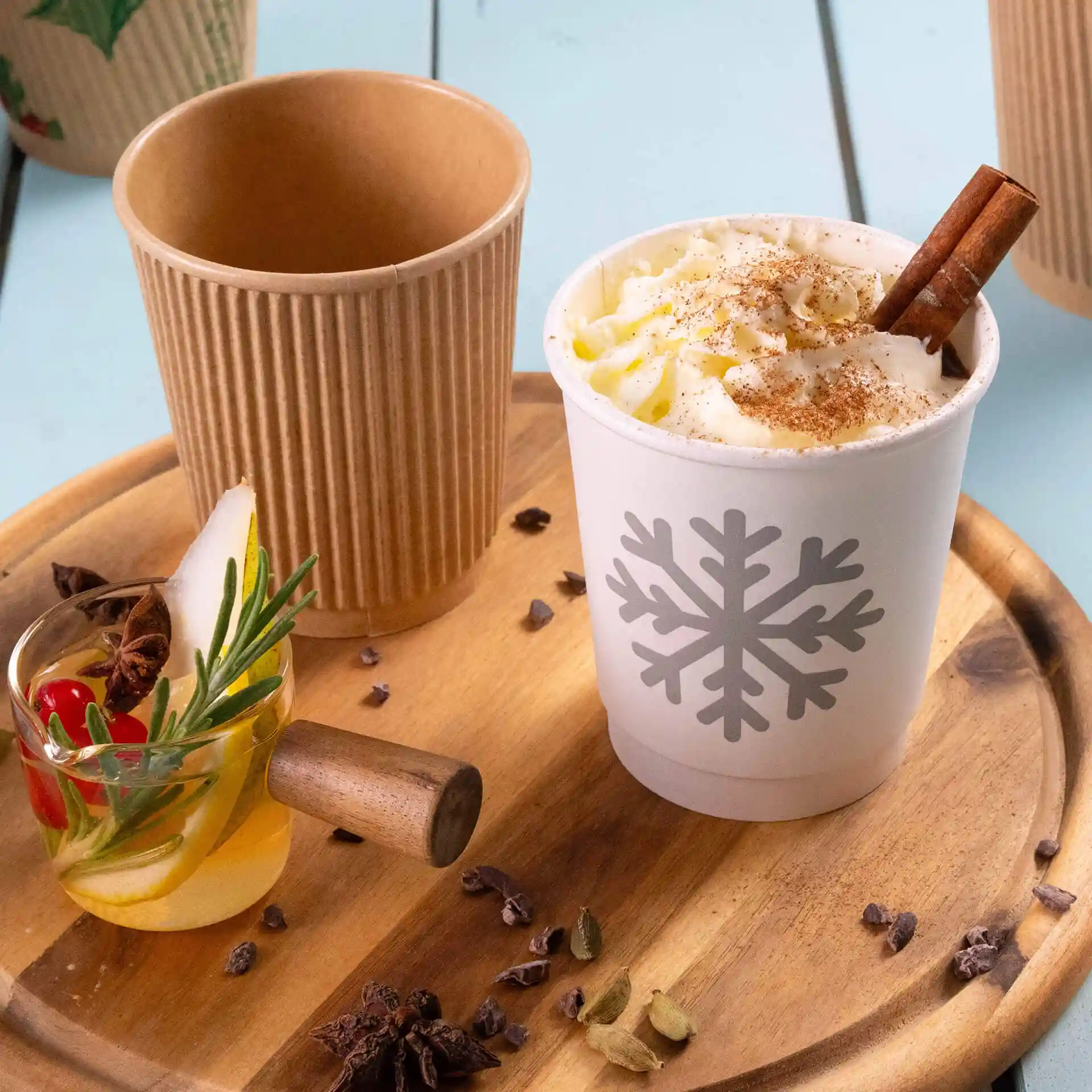 Gobelet blanc avec un design de flocon de neige, rempli de crème fouettée et d'un bâton de cannelle, à côté d'un gobelet kraft et d'une boisson garnie.