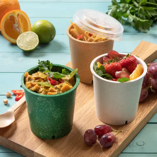 Trois tasses à soupe réutilisables sur une planche en bois, remplies de soupe, de fruits et de nouilles, entourées d'agrumes frais et de raisins.