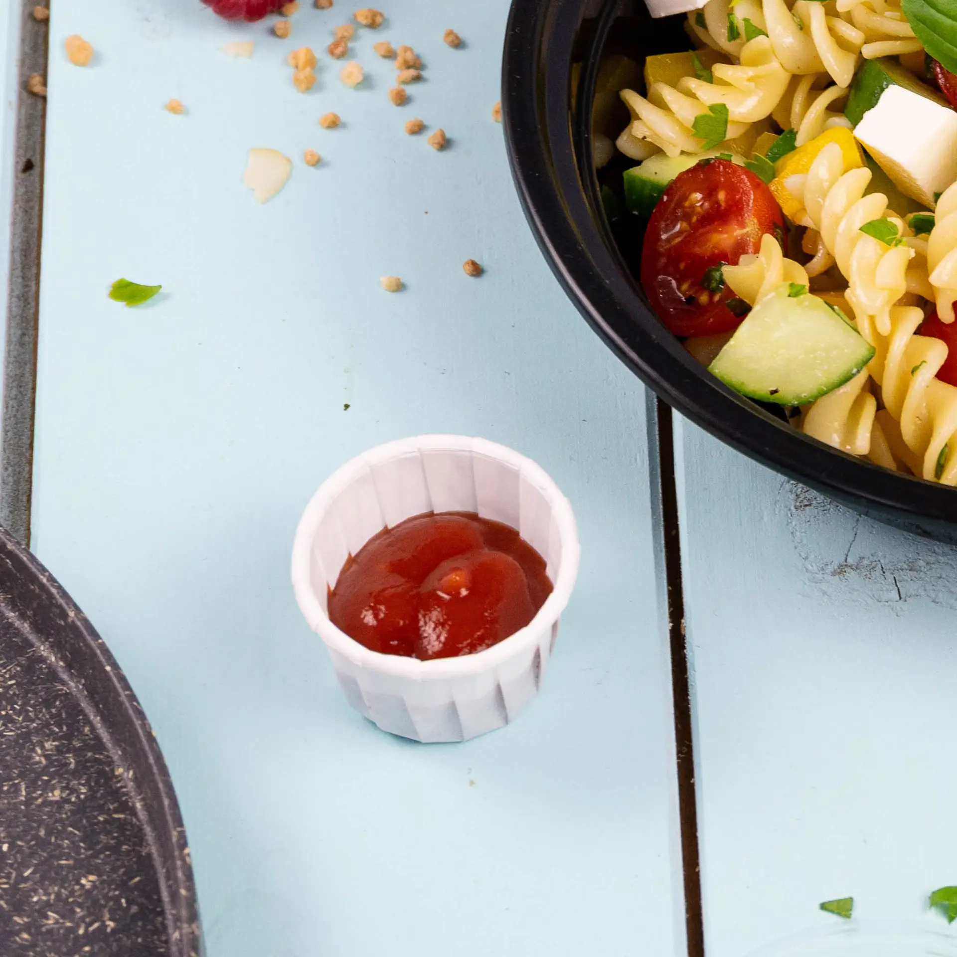 Petit gobelet en papier blanc rempli de ketchup, placé à côté d'un bol de salade de pâtes sur une surface en bois bleu.