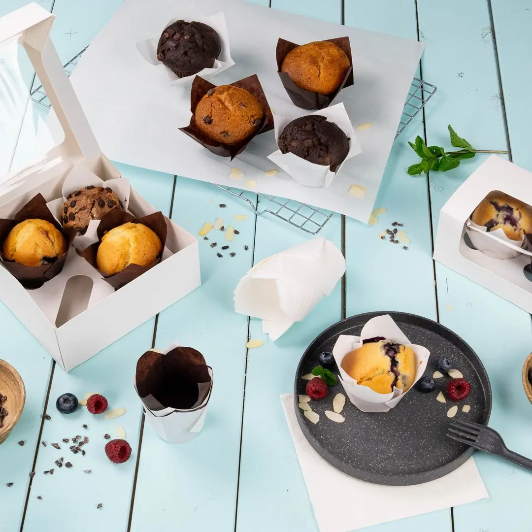 Muffins assortis dans des emballages en papier sur une surface bleu clair, avec une boîte blanche et une assiette de muffins et de baies.