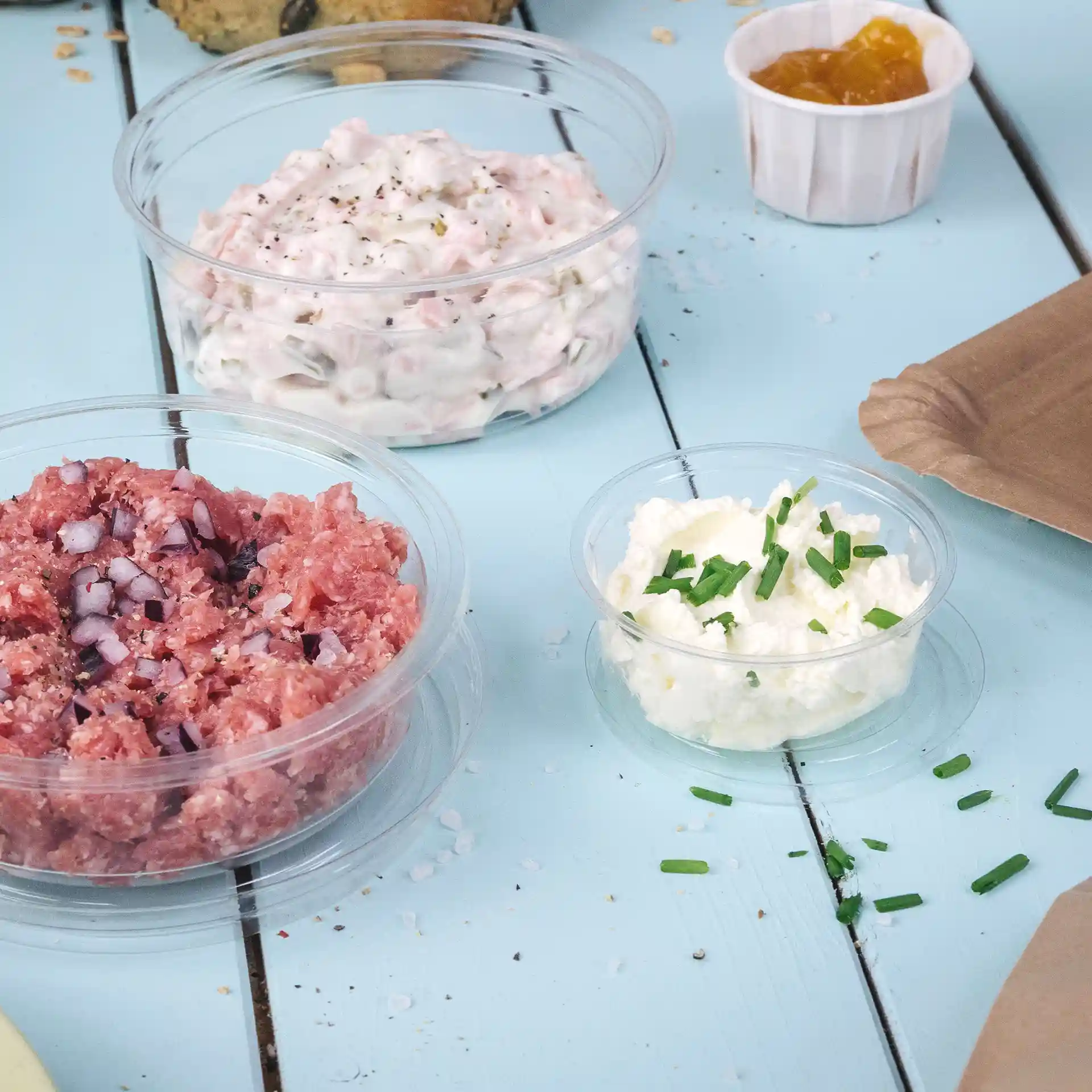 Trois gobelets en PLA transparents contenant différents aliments sur une surface en bois bleu.