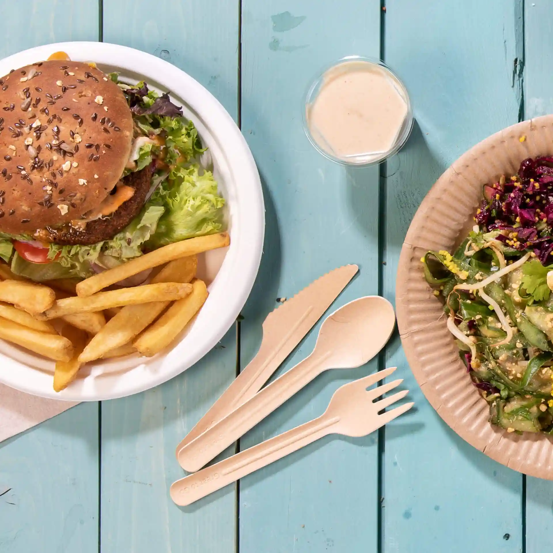 Image d'un burger avec de la laitue et des frites dans un bol blanc, à côté d'une salade dans une assiette en papier brun et un petit pot de sauce.