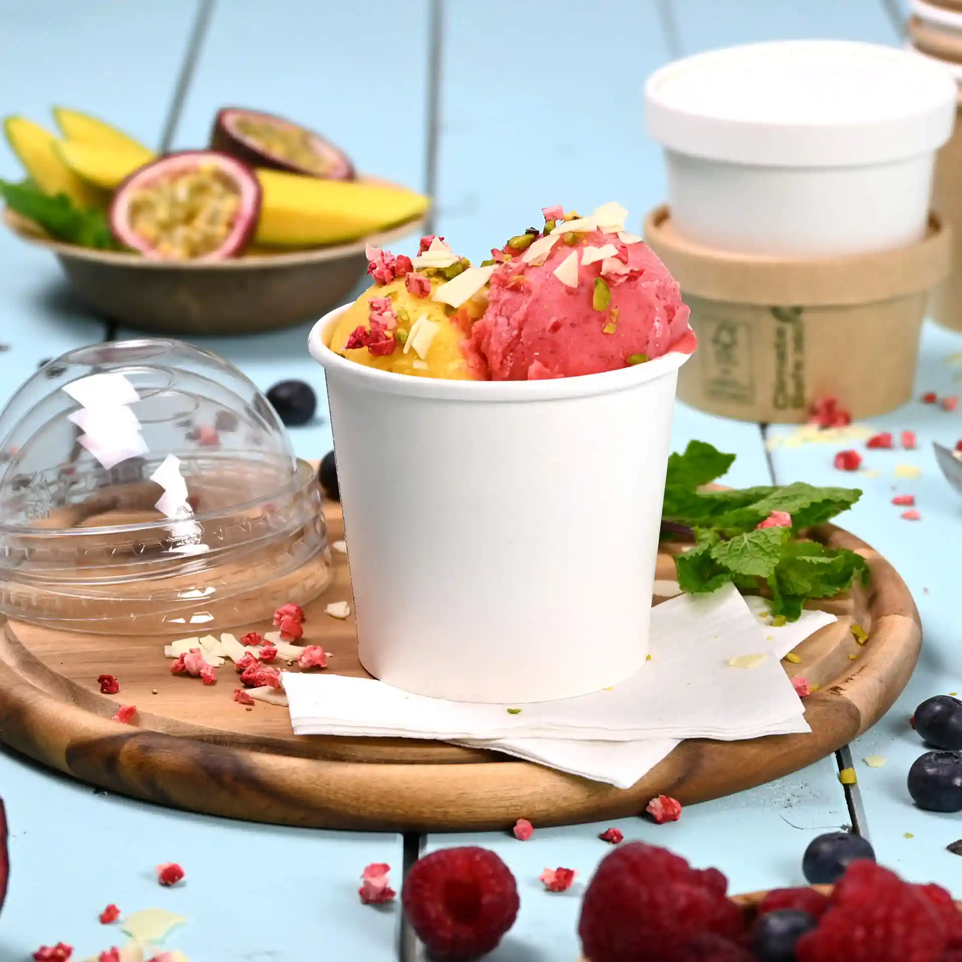 Tasse de crème glacée en papier blanc avec de la glace rose et jaune garnie de noix, placée sur un plateau en bois avec des fruits et des baies.